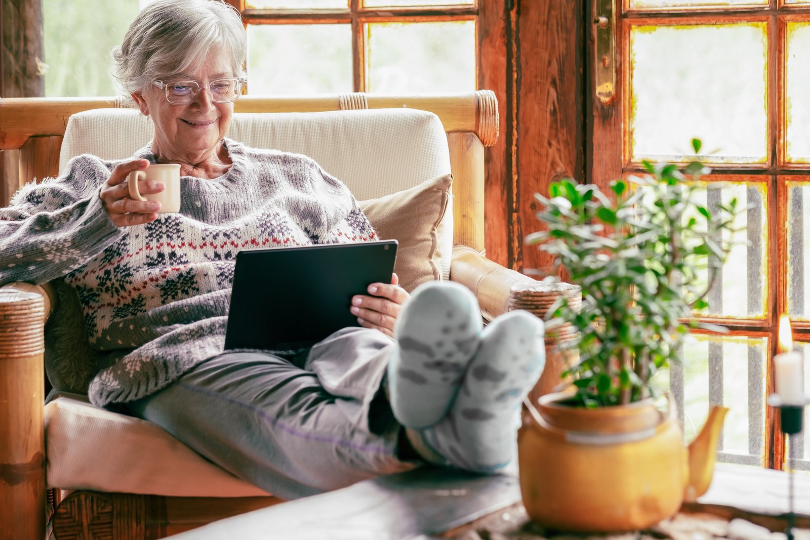 An older woman relaxing in a cozy rattan armchair with a cup of tea and a tablet, warm wooden windows and a potted plant beside her